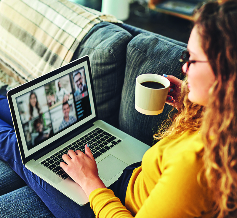 A woman attending an online meeting on a laptop whilst having coffee on the sofa at home